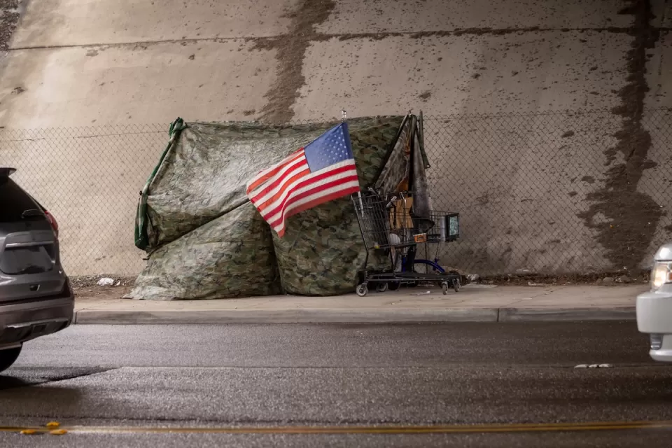 Tent covered with camouflage tarp with American flag on front under freeway overpass in California.